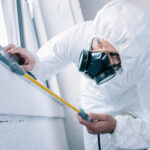 pest control worker in respirator spraying pesticides under windowsill at home